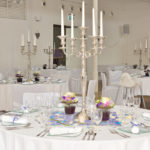 Blick auf viele runde Tische, weiß eingedeckt, Stühle mit weißen Hussen, Gläser und Teller am Tisch, 5-armige Kerzenleuchter